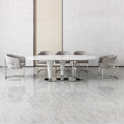 Cristallo dining room table with round white marble top and polished metal pedestal base, surrounded by grey velvet chairs.